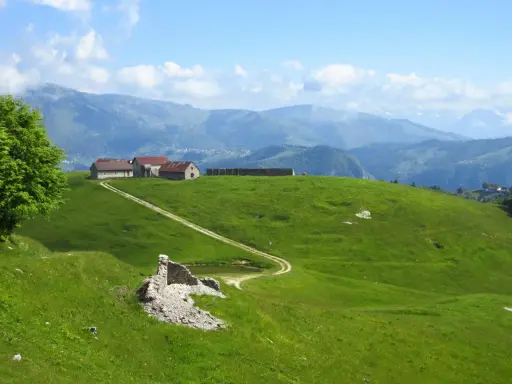 Un sentiero delle malghe serpeggia tra verdi colline, conduce a rustici edifici e si perde tra le montagne nebbiose.