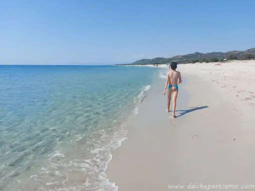 Un bambino cammina su una spiaggia di sabbia bianca, verso le spiagge più belle della Sardegna est. Acqua cristallina e cielo azzurro.