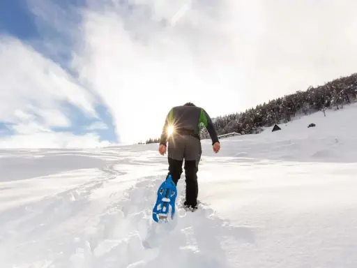 Uomo con ciaspole cammina su sentiero innevato, ideale per chi cerca dove ciaspolare in Piemonte. Sole splendente tra le nuvole.