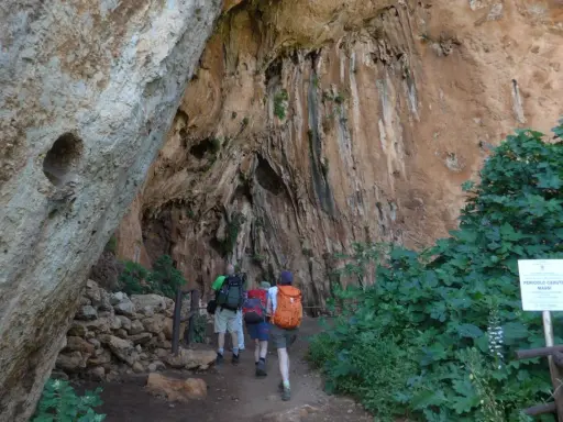 Escursionisti esplorano un sentiero nella Riserva dello Zingaro, avvicinandosi a un'imponente parete rocciosa con vegetazione rigogliosa.