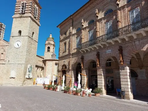 Piazza a San Ginesio, Marche, con il campanile in pietra e l'edificio in mattoni rossi con portici.