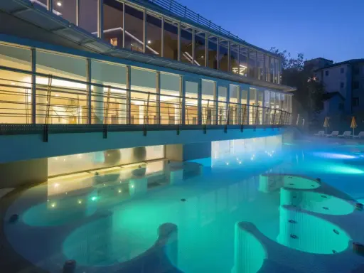 Piscina termale illuminata di notte, con vapori che salgono. Un edificio moderno con ampie vetrate si affaccia sull'acqua turchese, evocando un'atmosfera di relax e benessere, come in antiche terme preistoriche a Padova.