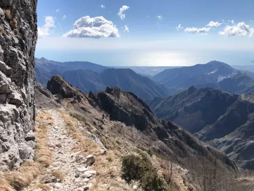 Sentiero roccioso su un crinale montuoso, con vista sul mare e sulle colline. Un percorso che ricorda la via Vandelli, un'antica strada panoramica.