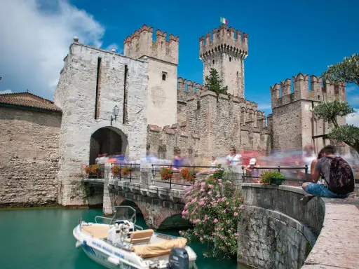 Castello Scaligero di Sirmione, un'ottima idea per cosa vedere a Sirmione in un giorno. Un ponte pedonale affollato porta all'ingresso del castello, con un motoscafo ormeggiato nell'acqua turchese.
