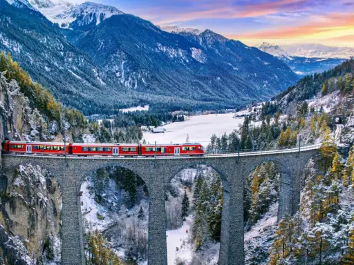 Il trenino del Bernina attraversa un viadotto in pietra in un paesaggio alpino innevato, con montagne maestose e un cielo colorato al tramonto.