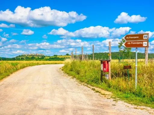 Segnaletica per la Via Francigena, un antico cammino. La strada sterrata conduce verso un borgo collinare sotto un cielo azzurro con nuvole bianche.