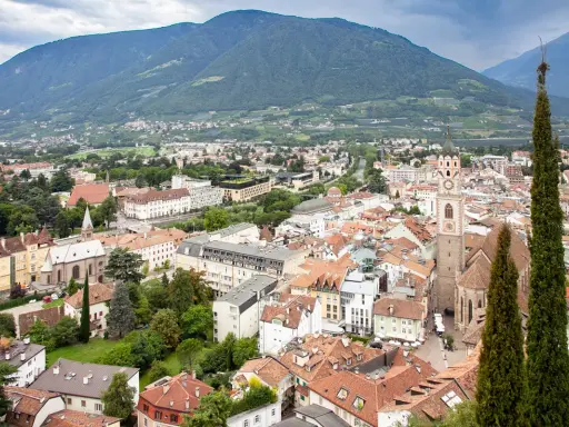 Vista aerea del centro storico di Merano, con montagne sullo sfondo e un campanile che svetta tra i tetti rossi.