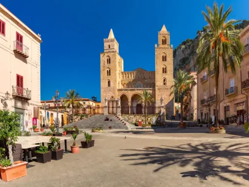 Piazza soleggiata con la cattedrale normanna sullo sfondo, palme e palazzi storici. Un luogo incantevole tra i borghi da visitare in Sicilia.