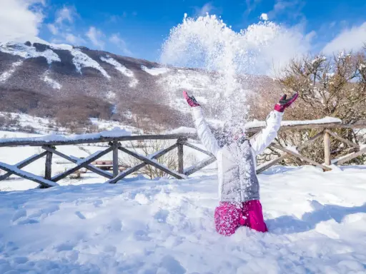 Bambina felice lancia neve in aria, con montagne innevate sullo sfondo. Perfetto per scoprire dove andare per la neve in Abruzzo.