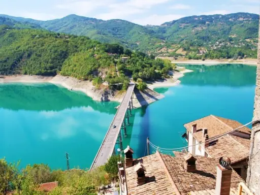Panoramica mozzafiato del lago turchese vicino a Castel di Tora, con un ponte che attraversa le acque cristalline e montagne verdi sullo sfondo.