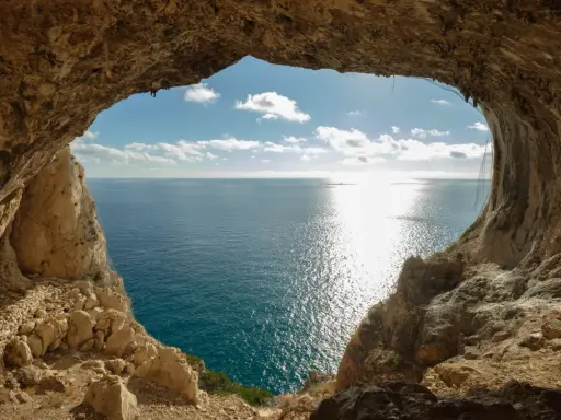 Vista dal mare da una grotta, con un sentiero del pellegrino che si intravede tra le rocce. Il sole brilla sull'acqua.