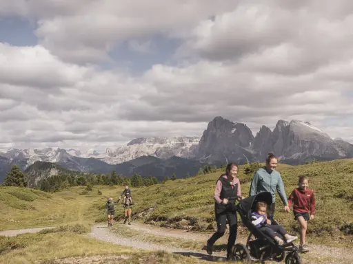 Famiglia in escursione sull'Alpe di Siusi, con bambini e passeggino, sullo sfondo le maestose vette dolomitiche.