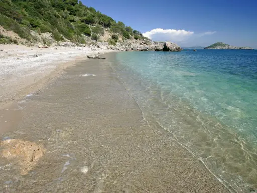 Spiaggia di ciottoli e sabbia con acqua cristallina, vista verso il mare aperto e un'isola in lontananza, vicino al porto di Ercole.