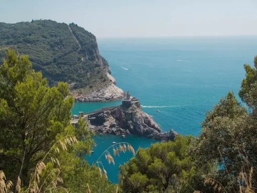 Vista panoramica del sentiero Riomaggiore Portovenere, con la chiesa di San Pietro su uno sperone roccioso e il mare azzurro.