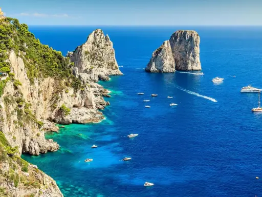 Scogliere verdi e rocciose si affacciano sul blu intenso del golfo di Napoli, punteggiato da barche e yacht. Faraglioni iconici emergono dal mare.