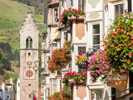 Torre medievale con orologio e case fiorite, un'immagine che mostra dove si trova Vipiteno, con verdi colline sullo sfondo.