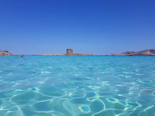 Acque cristalline del mare nord Italia con torre antica su isolotto e barca a vela arancione.