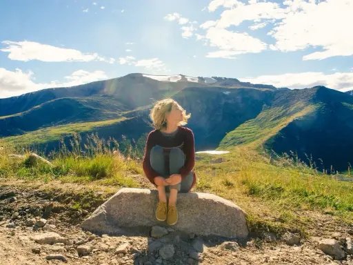 Ragazza seduta su una roccia, con i capelli mossi dal vento, ammira il panorama montano. Un momento di pace mentre camminare nella natura.