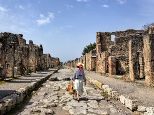 Una donna con cappello di paglia cammina lungo una strada lastricata di Pompei, tra antiche rovine. Un'esperienza unica per scoprire cosa vedere a Pompei.