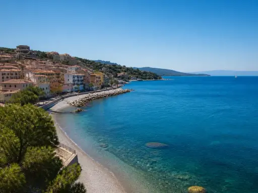 Porto Santo Stefano: case colorate affacciate su spiagge di ciottoli e sabbia, con il mare azzurro e un veliero all'orizzonte.