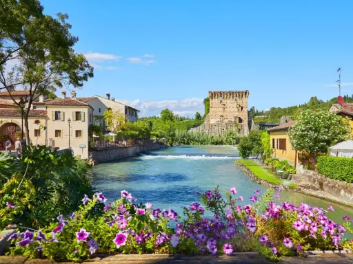 Un fiume attraversa un pittoresco borgo medievale veneto, con un'antica torre in rovina e case colorate.