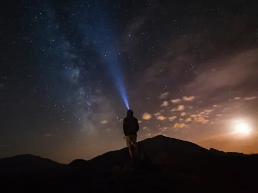 Una persona con una torcia illumina il cielo stellato su una montagna di notte. La Via Lattea è visibile.