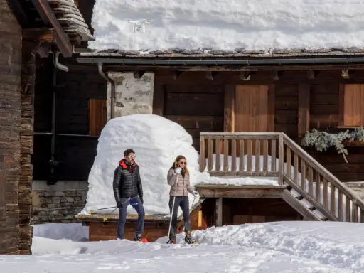 Coppia con racchette da neve a Riale Val Formazza, con un drone in volo sopra un tetto innevato.