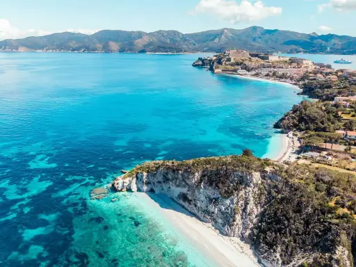 Costa rocciosa con spiaggia bianca e mare cristallino, un borgo fortificato domina la baia. Splendida località mare Toscana.