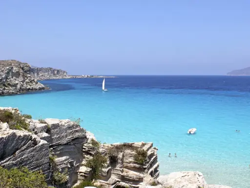 Scogliere rocciose e mare cristallino a Favignana. Un veliero solca le acque turchesi, perfetto per scoprire cosa vedere a Favignana in un giorno.