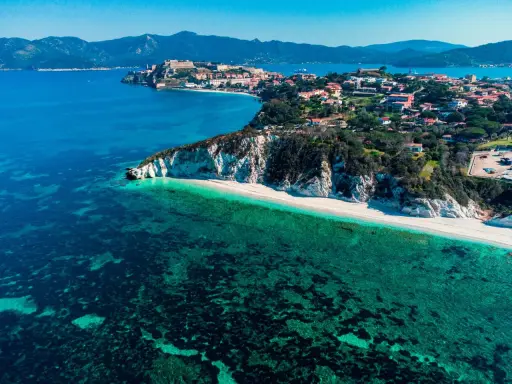 Vista aerea di una spiaggia bianca con acque cristalline e un borgo storico su una collina, parte dell'arcipelago toscano isole visitabili.