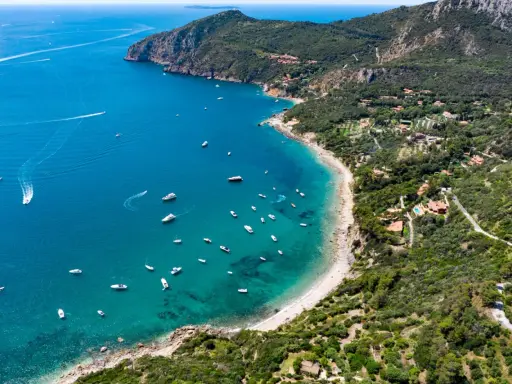 Baia con acque cristalline e numerose barche, incastonata tra colline verdi e scogliere. Un vero gioiello tra le spiagge più belle Toscana.
