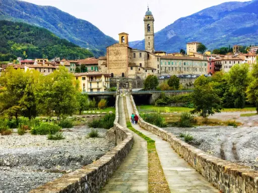 Ponte Gobbo e borghi vicino a Bobbio, con montagne sullo sfondo. Due persone passeggiano sul ponte antico.