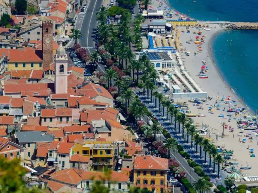 Aerial view of a coastal town with red-tiled roofs, a beach with umbrellas, and palm trees lining the road.