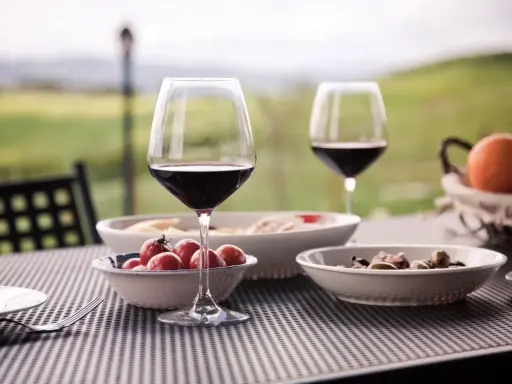 Un bicchiere di vino rosso e stuzzichini su un tavolo all'aperto, con un paesaggio collinare sullo sfondo, perfetto per un percorso enogastronomico toscana.