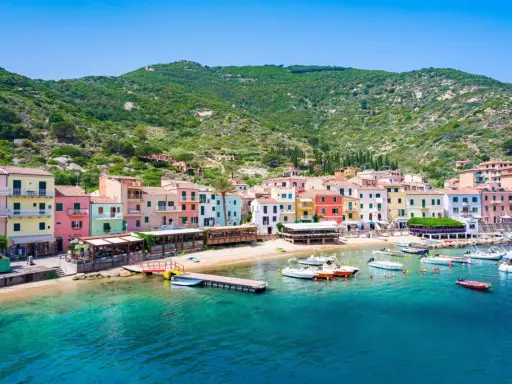 Colorate case affacciate sul mare cristallino, con barche ormeggiate e la collina verde sullo sfondo. Un'immagine che evoca il fascino del traghetto per l'Isola del Giglio.
