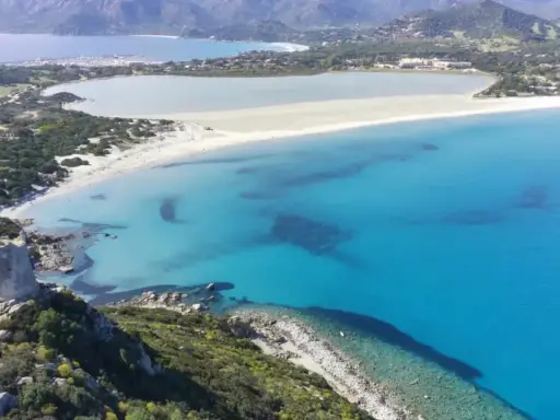 Torre costiera su promontorio verdeggiante domina una baia con acque cristalline, tra le spiagge più belle Sardegna.