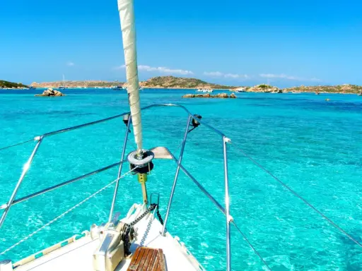La prua di una barca a vela naviga verso La Maddalena, tra acque cristalline e isole rocciose.