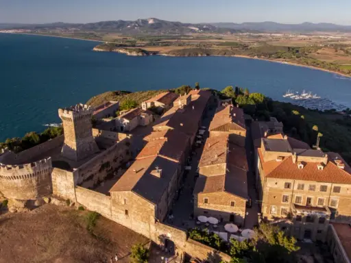 Borgo medievale fortificato domina la campagna toscana lungo il mare, con un castello imponente e case dai tetti rossi affacciate su un golfo con barche a vela.