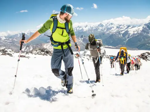 Un gruppo di escursionisti sta camminando sulla neve in montagna, con bastoncini da trekking e zaini, sotto un cielo azzurro.