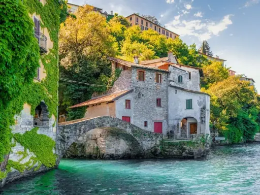Antichi borghi medievali lombardia da visitare: un ponte di pietra collega case storiche sull'acqua cristallina, con edera rampicante e vegetazione lussureggiante.