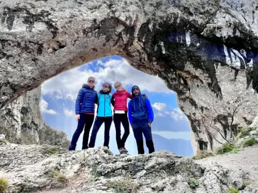 Quattro amici si godono una giornata di camminare in montagna, posando sotto un arco naturale di roccia con un cielo azzurro e nuvoloso.