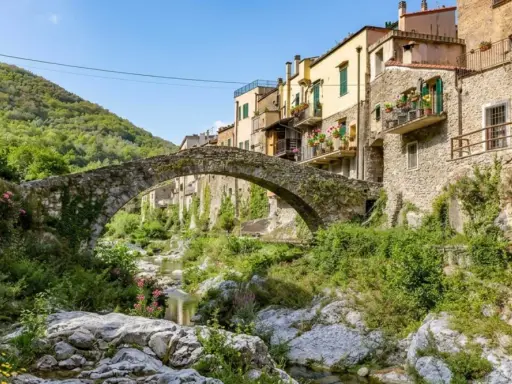 Antico ponte in pietra collega i borghi dell'entroterra ligure ponente, con case colorate e vegetazione rigogliosa che scende verso il torrente.