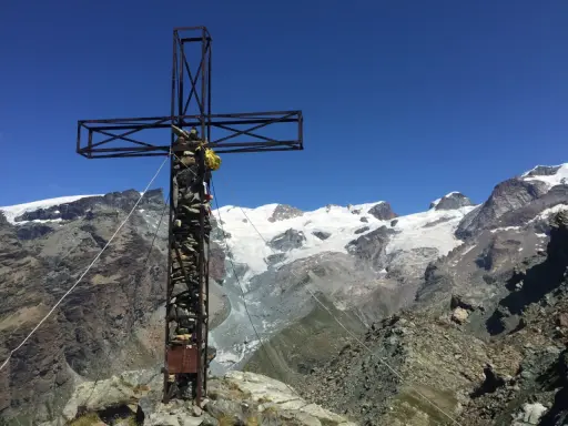 Croce metallica e di pietra in cima a una montagna, con un ghiacciaio e vette innevate sullo sfondo. Il **palon di Resy** domina il panorama.