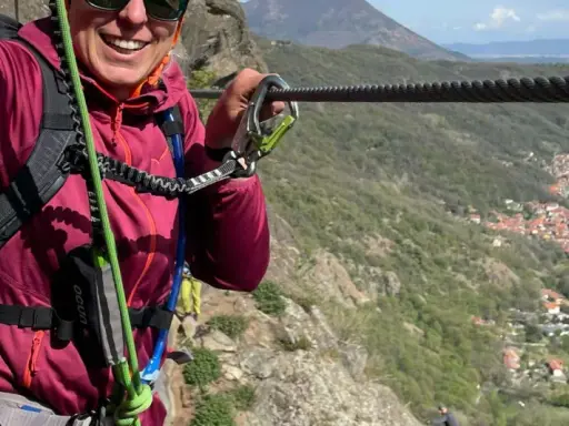 Donna sorridente si aggrappa a un cavo d'acciaio, godendosi un'avventura sulle ferrate Val di Susa con un panorama mozzafiato.