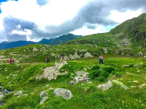 Escursionisti attraversano un paesaggio verdeggiante in una località montagna Piemonte, con rocce e nuvole minacciose all'orizzonte.
