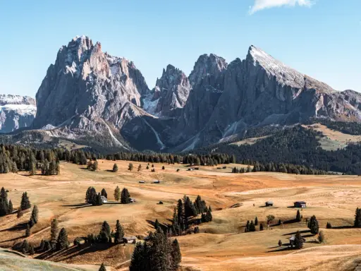 Un paesaggio mozzafiato delle Dolomiti più belle, con cime imponenti, prati dorati e boschi verdi. Piccole baite sparse creano un'atmosfera idilliaca.