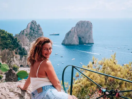 Donna sorridente con vista sui Faraglioni, un'ispirazione per come visitare Capri. Mare azzurro e barche in lontananza.