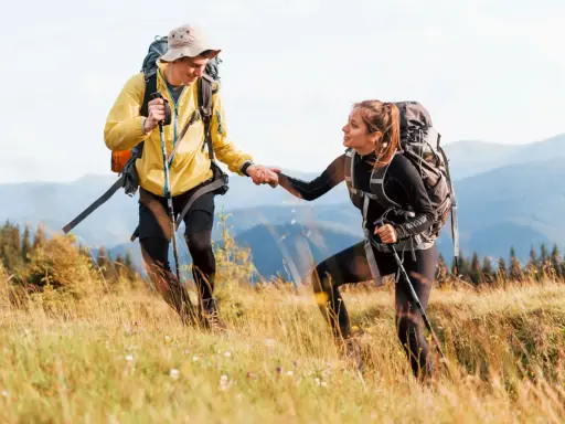 Coppia di escursionisti si aiutano a vicenda su un sentiero di montagna, con zaini e bastoncini da trekking.
