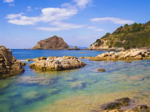 Acque cristalline e rocce scolpite dal mare a Porto Ercole, con montagne verdi sullo sfondo e un cielo azzurro punteggiato di nuvole.
