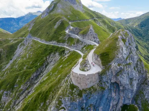 Un tortuoso cammino del sale si snoda tra montagne verdi, con ciclisti che affrontano tornanti mozzafiato su un sentiero panoramico.